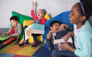 Four kids sitting on colorful bean bags, laughing and smiling with books in their hands.