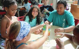 A group of four kids play Jenga on a table with an adult male volunteer. The child in the center of the photo is smiling at the camera.