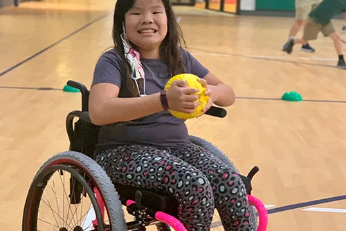 Young girl smiling in wheelchair, holding yellow ball in basketball gym