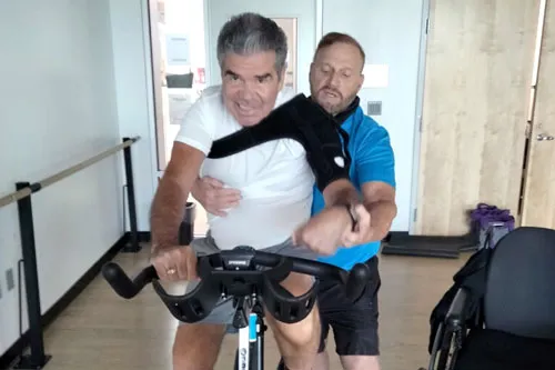 Personal trainer aids stroke victim on a spin bike.