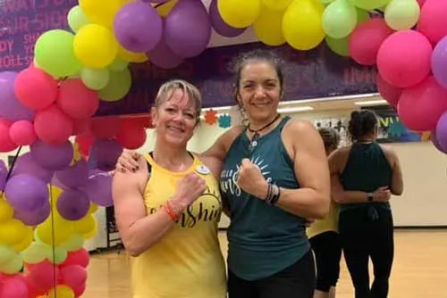 Group exercise instructor and Y member side hug while flexing biceps. The two pose for the photo after Les Mills Launch, under a balloon arch.