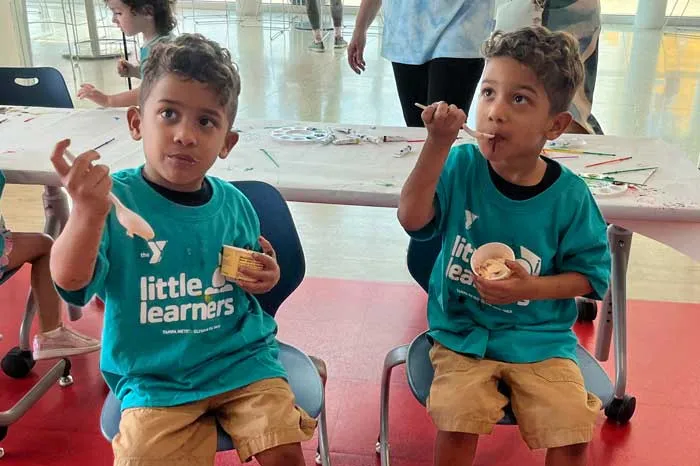 Two young children eating ice cream in the Spurlino Family Y's Little Learner's program. The pair are indoors, wearing teal shirts with the program name, khaki shorts and looking up from their treat.