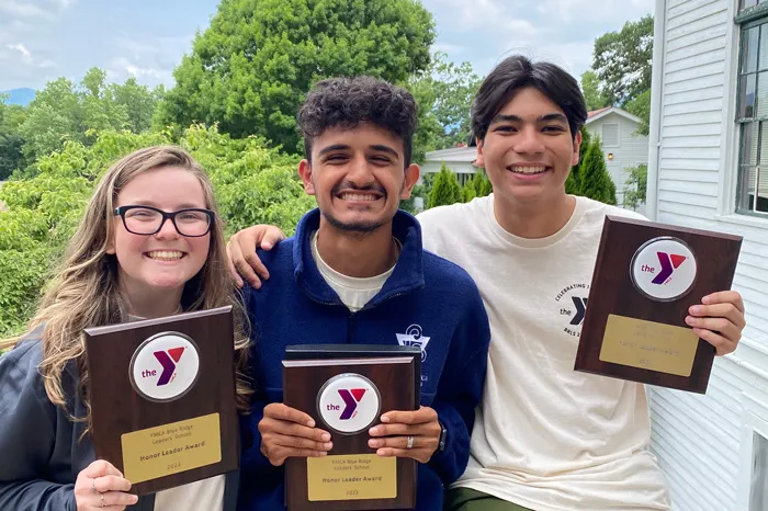 Three Tampa Y teen leaders holding their YMCA Honor Leader plaques. Green trees and cabins in the background.