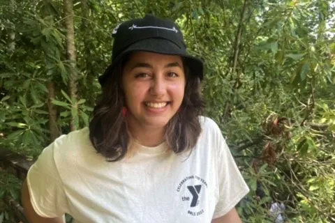 Teen wearing a white YMCA t-shirt and black bucket hat. Green shrubs and trees in the background.