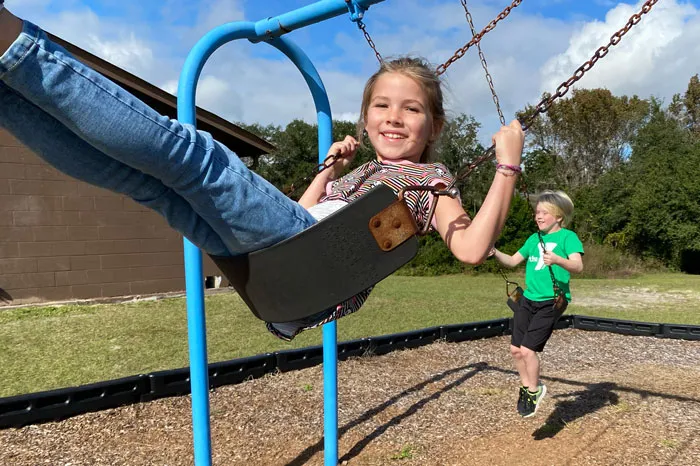 two kids on outdoor swings. girl wearing jeans in the center image, boy wearing green shirt swings in background.