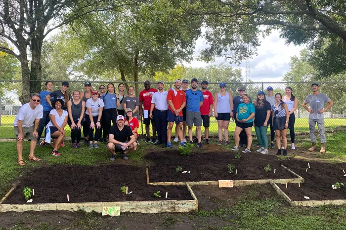 Volunteer group of 26 adults stand behind large raised garden beds outdoors. Trees and chainlink fence in the background.