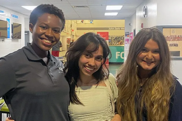 Three women wearing light green and gray shirts smile and pose together for a photo.