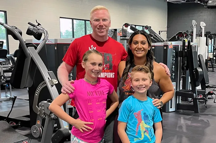 Family of four (Dad, mom, two children) pose together for a photo in the middle of the Northwest YMCA Wellness Floor. 