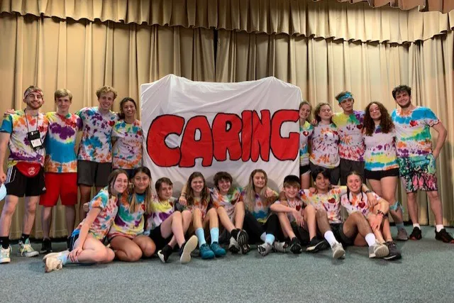 Group of teens wearing tie dye shirts gathered with a large DIY Caring sign. Sign has a white background and large, filled in red letters for "Caring."
