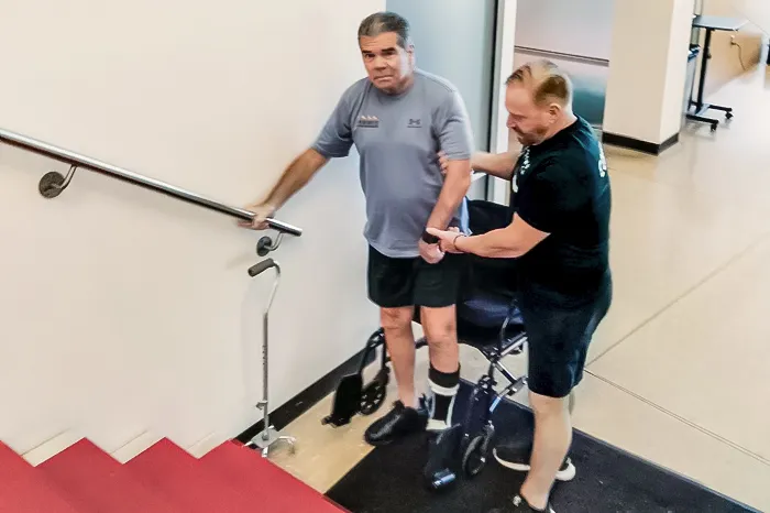 YMCA personal trainer and Y member regaining strength after a stroke, stand at the bottom of a staircase. The member stands in front of his wheelchair and holds onto the railing, while the trainer supports him.