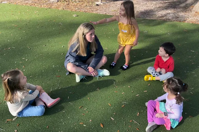 Camp Director sitting in circle on green turf with three young kids. One child is standing up playing the game duck duck goose.