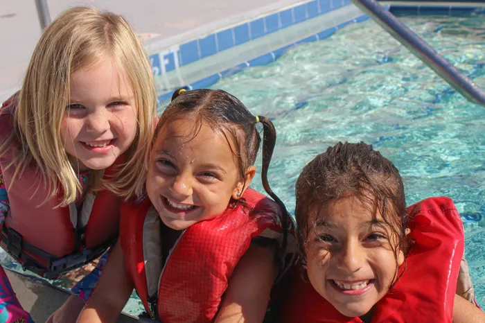 Three kids smiling and wearing lifejackets at entrance of pool.