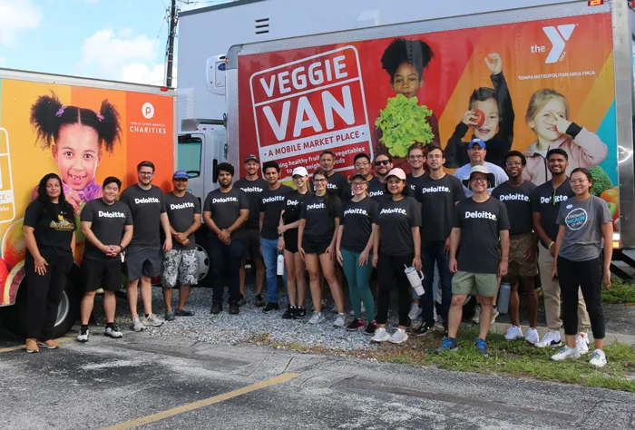 A group of 20 volunteers from Deloitte, wearing black t-shirts with their white logo, stand for a photo in front of Tampa YMCA's Veggie Van. The scene is outdoors on a sunny day. The group stands in a parking lot and patches of rocks and grass. 