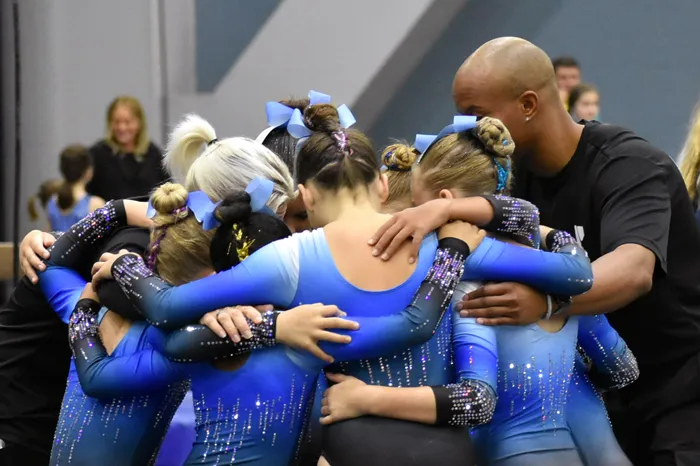 North Brandon Top Flight gymnastics team huddles with their coach at the 2023 YMCA National Gymnastics Championship. The coach wears a black short sleeve shirt and youth gymnasts wear blue and black gradient performance leotards with vertical line sparkles.