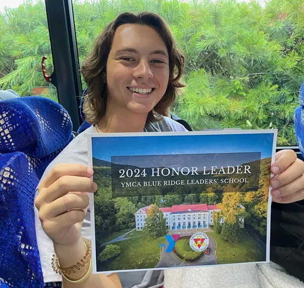 Teen smiles while holding a paper that reads 2024 Honor Leader YMCA Blue Ridge Leaders' School. The teen is on a bus with part of the blue seat showing, and luscious greenery outside. 