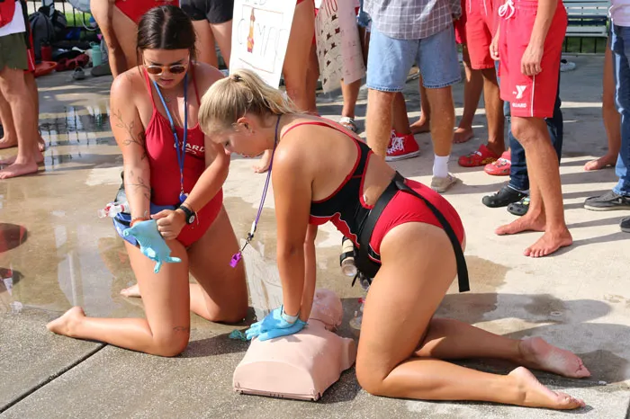 Two lifeguards training CPR with practice torso on an outdoor pool deck. Background: legs of other lifeguards watching the drill.
