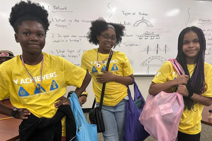 Three female teens wearing yellow YMCA Achievers shirts hold new bags filled with hygiene and period products.