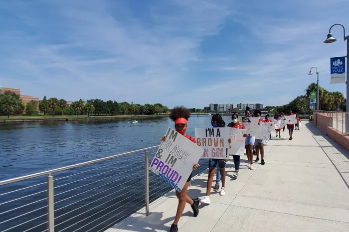 Tampa YMCA Teen Achievers march along the Riverwalk on a sunny day. The teens are lined up, single file, each holding a sign about their personal pride and identity.