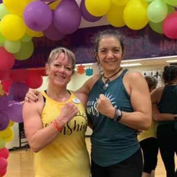 Group exercise instructor and Y member side hug while flexing biceps. The two pose for the photo after Les Mills Launch, under a balloon arch.