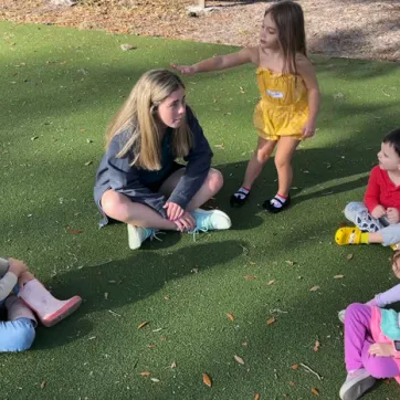 Camp Director sitting in circle on green turf with three young kids. One child is standing up playing the game duck duck goose.