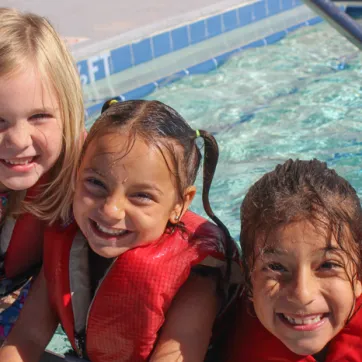 Three kids smiling and wearing lifejackets at entrance of pool.