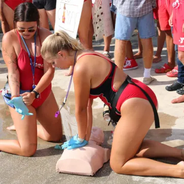 Two lifeguards training CPR with practice torso on an outdoor pool deck. Background: legs of other lifeguards watching the drill.