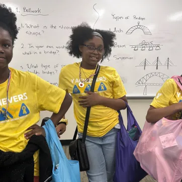 Three female teens wearing yellow YMCA Achievers shirts hold new bags filled with hygiene and period products.