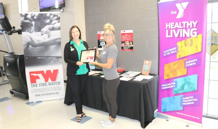 photo taken with two women - one receiving a Fire Watch certificate from the other in between two banners - one for Fire Watch and one for YMCA Healthy Living initiatives.